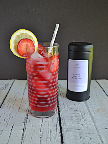 A glass of berry iced tea with a lemon slice and strawberry slice and a straw in the glass with a container of tea behind the glass.