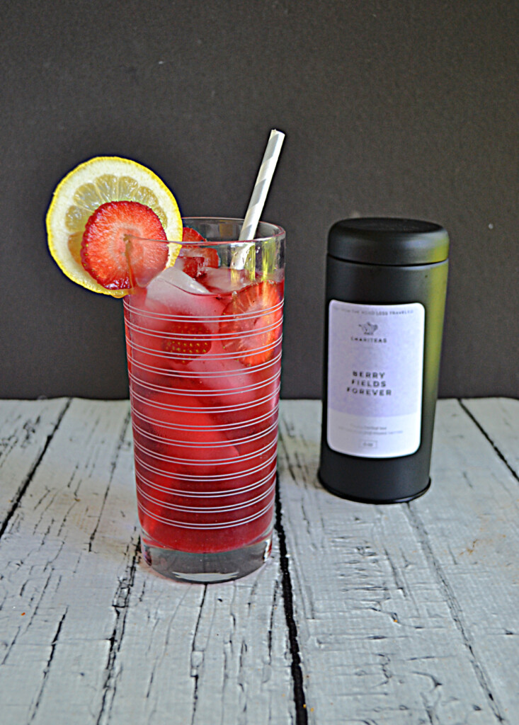 A glass of berry iced tea with a lemon slice and strawberry slice and a straw in the glass with a container of tea behind the glass. 