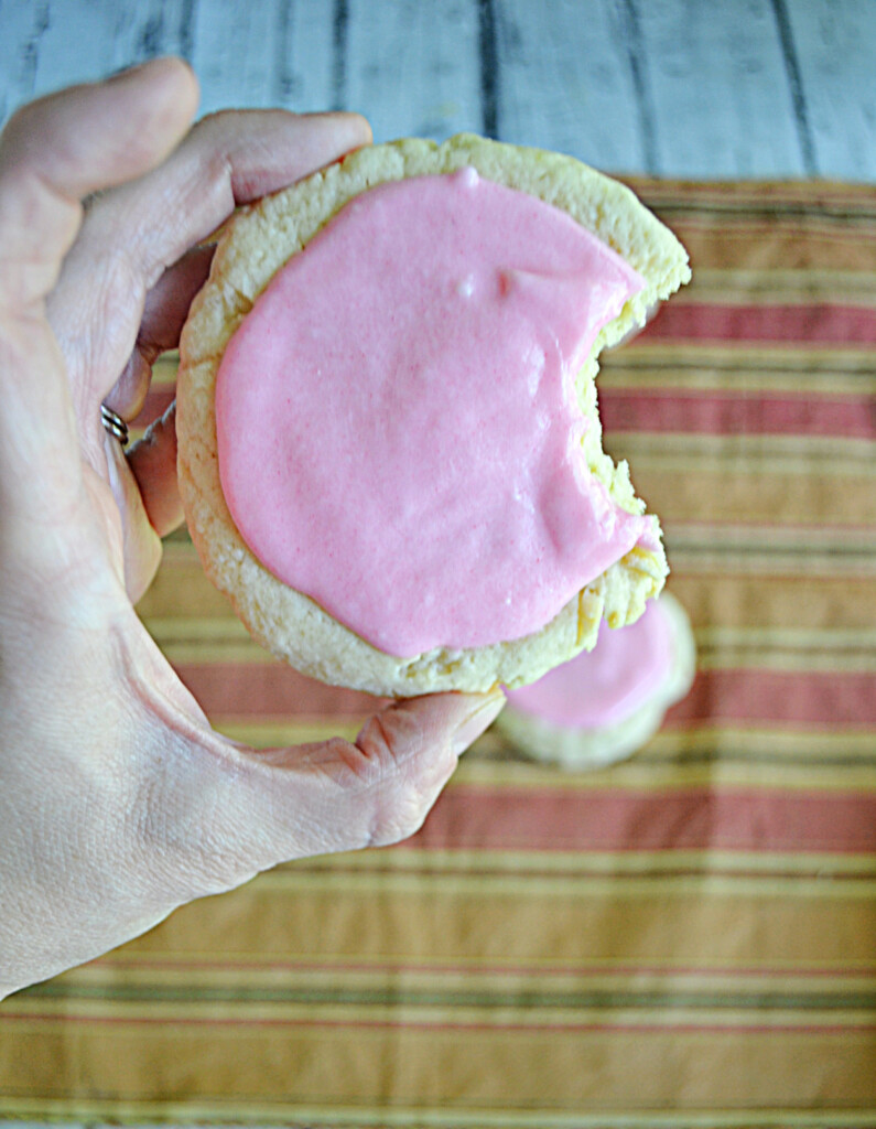 A front view of a hand holding a sugar cookie with pink frosting and a bite taken out of it.