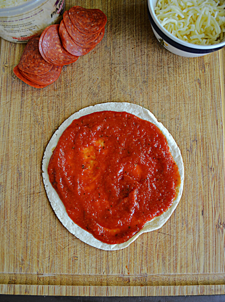 A cutting board with a tortilla topped with pizza sauce. 