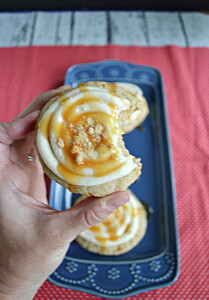 A close up of a large cookie with cream cheese frosting, a caramel drizzle, and graham cracker crumbs on top with a big bite taken out of it.