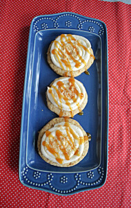 A top view of a blue platter with three large cookies topped with cream cheese frosting, a caramel drizzle, and graham cracker crumbs on top.