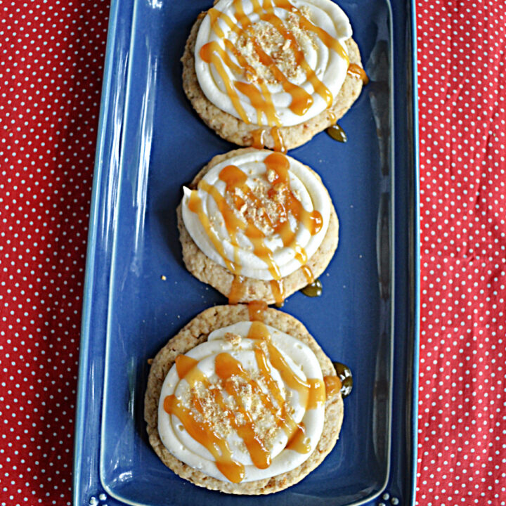 A top view of a blue platter with three large cookies topped with cream cheese frosting, a caramel drizzle, and graham cracker crumbs on top.
