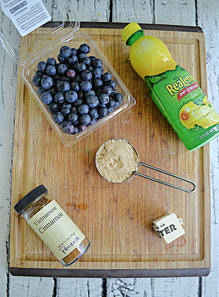 A cutting board with blueberries, a bottle of lemon juice, brown sugar, a stick of butter, and a jar of cinnamon on it.