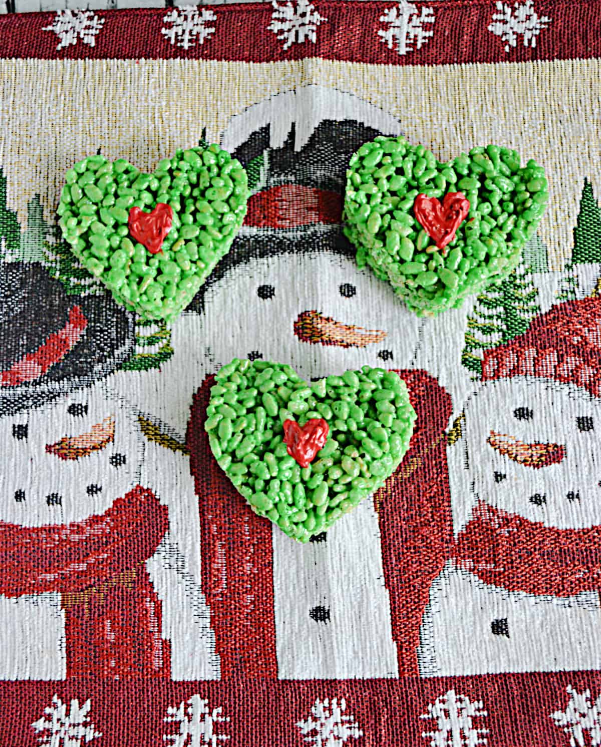 Three green heart shaped Rice Krispies Treats with a red heart in the middle on a snowman background. 