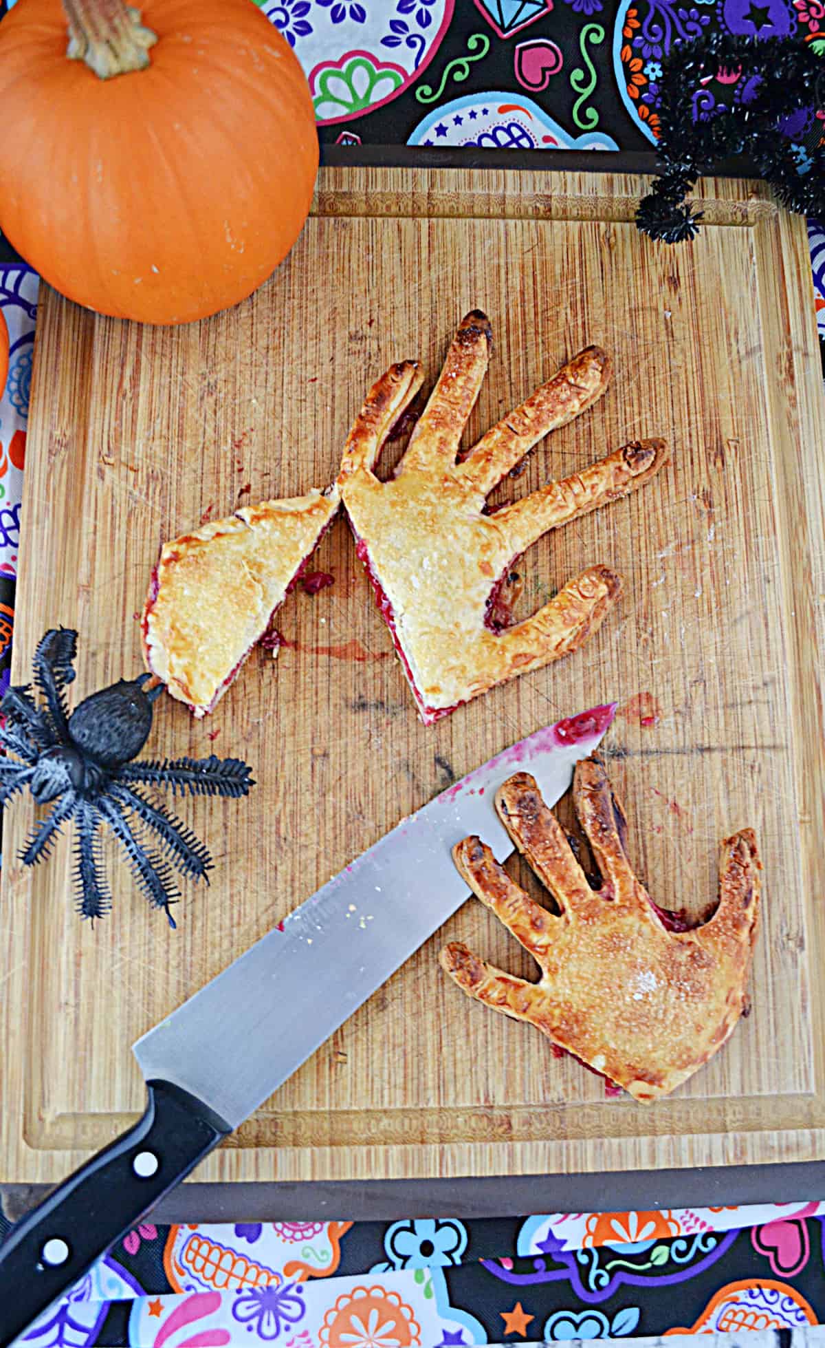 Two severed hand pies with a spider crawling towards them, a knife on the board, and a pumpkin behind it.