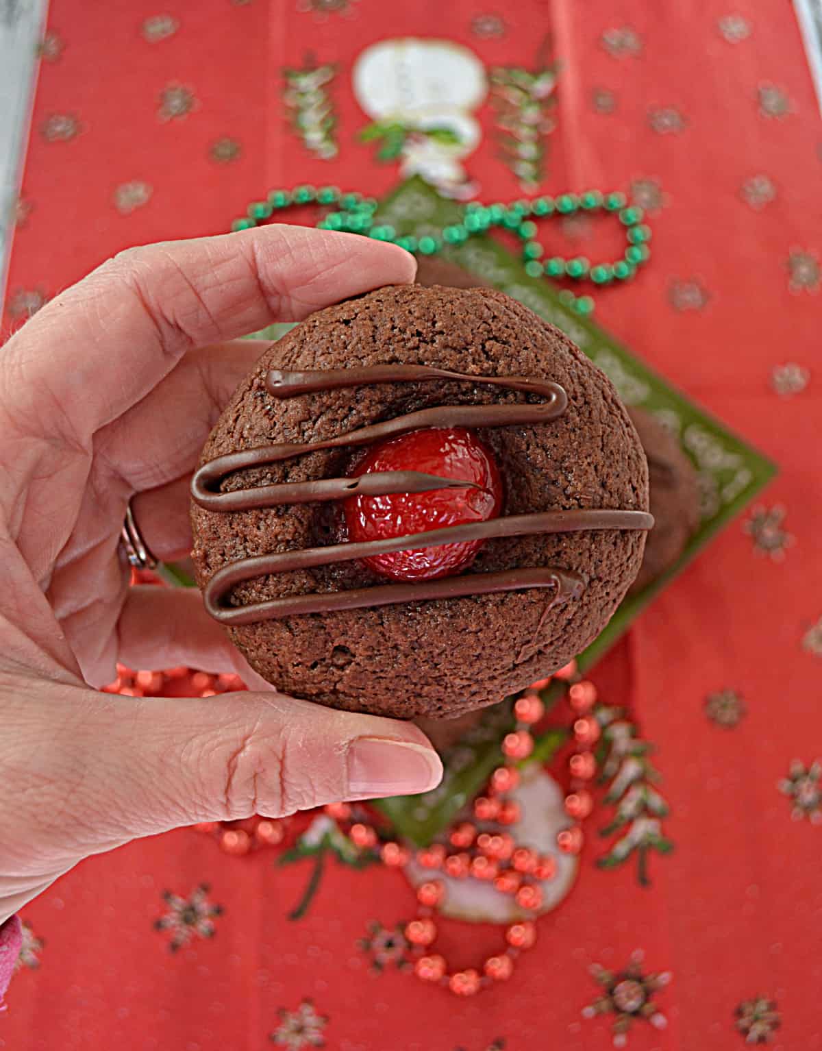 A close up of a hand holding a chocolate covered cherry thumbprint cookie. 