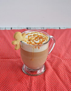 A mug of Gingerbread Latte with a caramel drizzle and a gingerbread man on the rim.