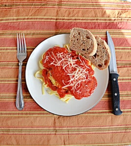 A plate of Chicken Parmesan with 2 pieces of bread on the plate and a knife and fork next to the plate.