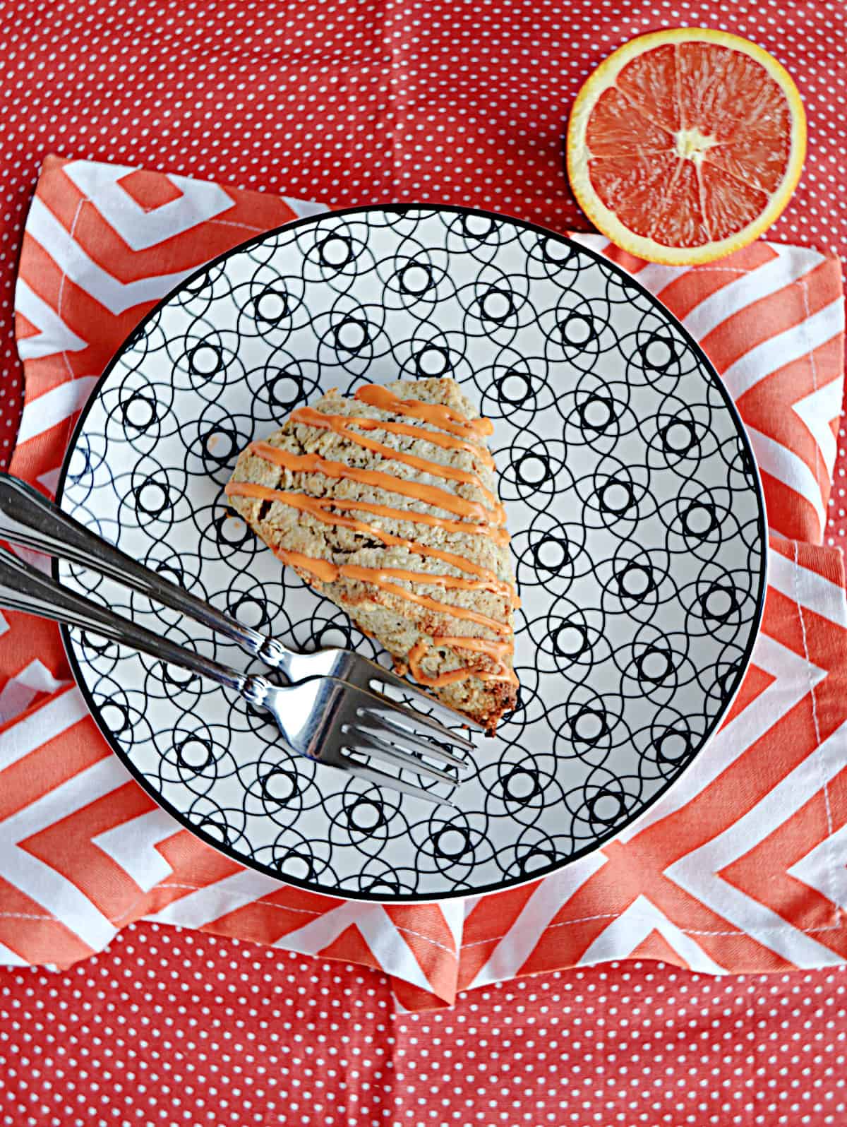 A plate with a scones drizzled with glaze, 2 forks on the plate, and an orange in the background. 