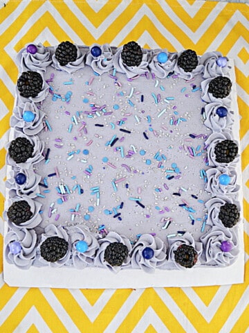 A purple, square cake topped off with sprinkles and fresh blackberries.