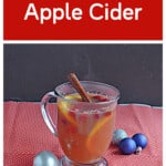 Pin Image: Text title, a mug of cider with a cinnamon stick and orange slices with blue and silver ornaments around it.
