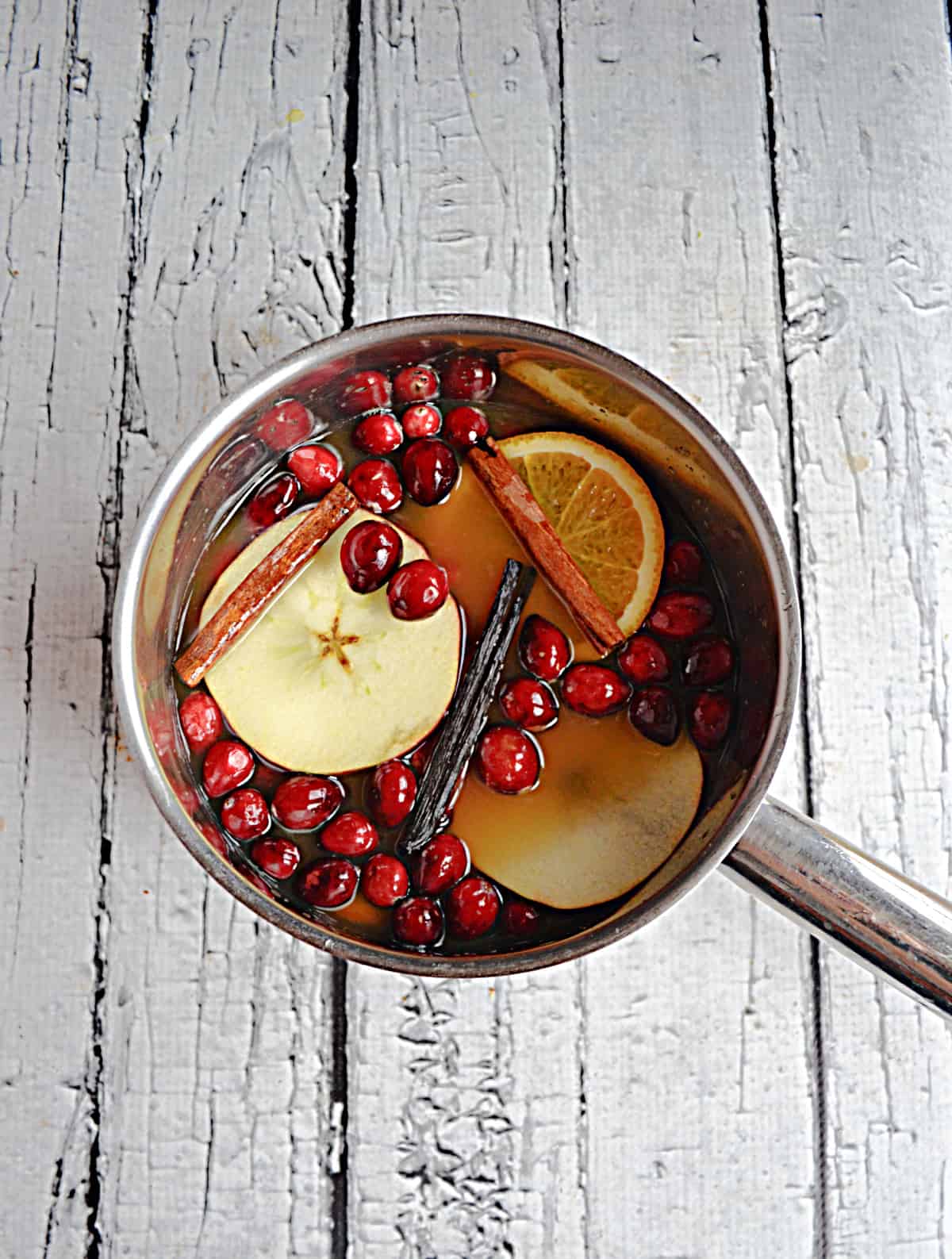 A saucepan with apple cider, sliced apples, fresh cranberries, and cinnamon sticks in it.