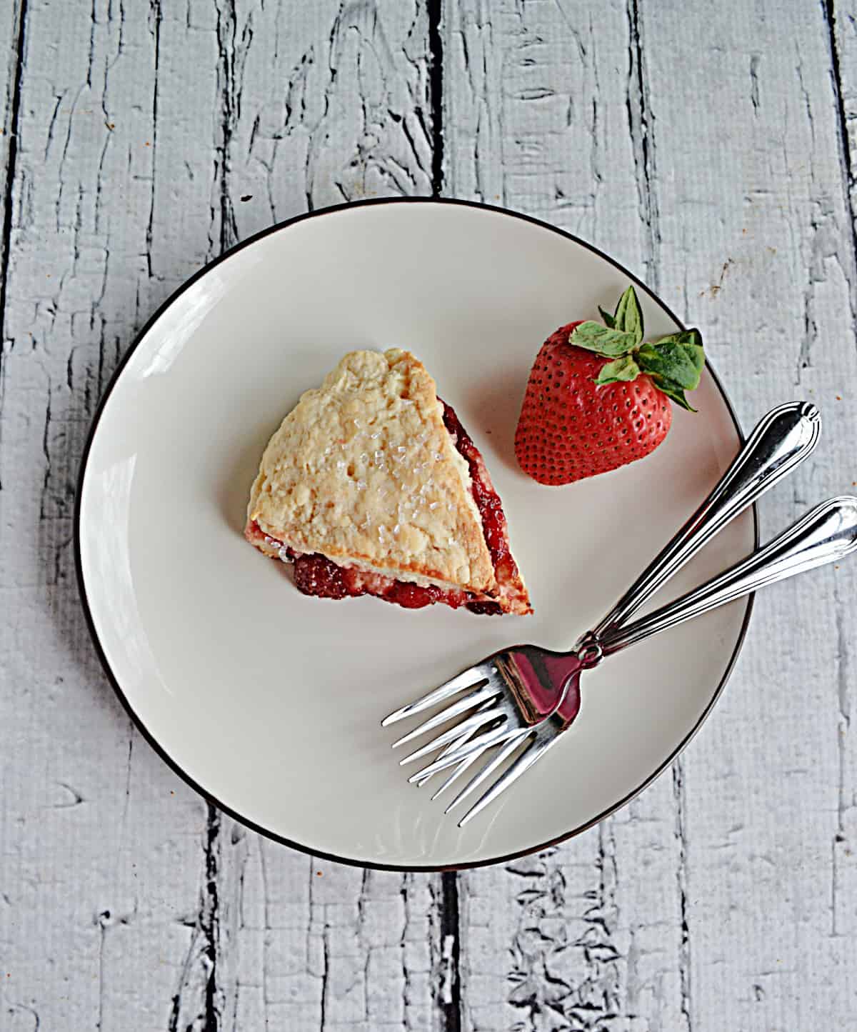 A plate with a scone on it with two forks and a fresh strawberry.