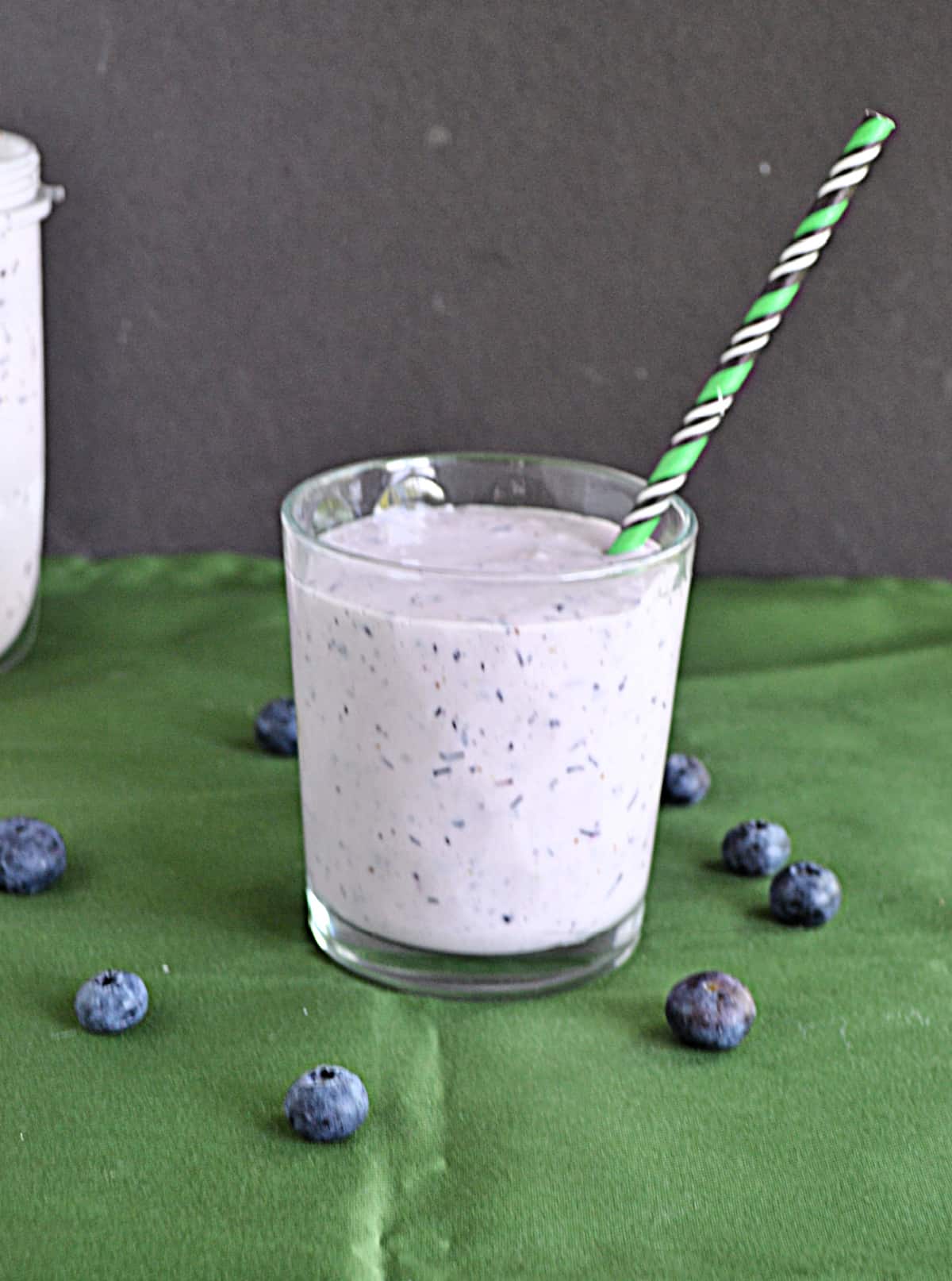 A blueberry banana smoothie in a glass with a straw and blueberries around the glass. 