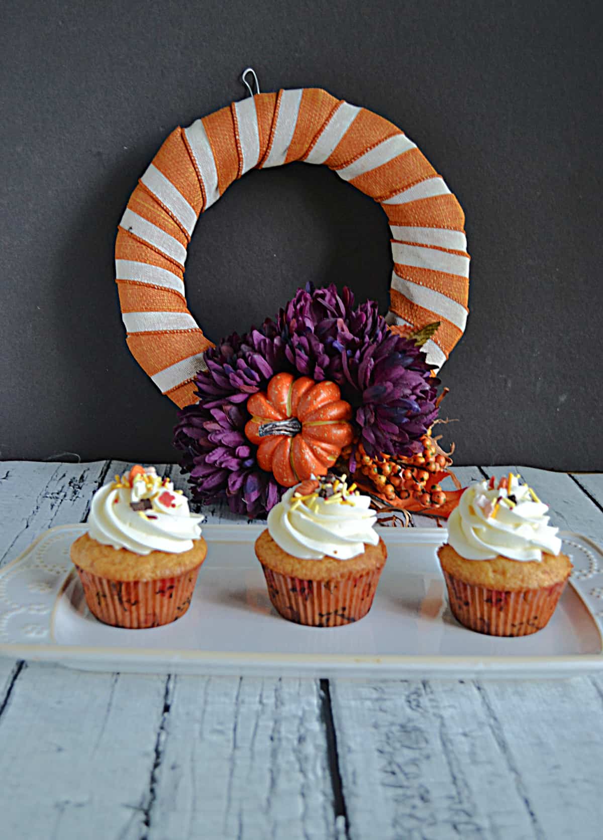 An orange and white fall wreath with three cupcakes in front of it topped with frosting and fall colored sprinkles. 
