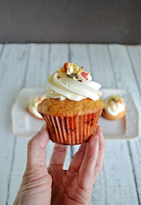 A close up of a hand holding a butterscotch cupcake with frosting and fall colored sprinkles.