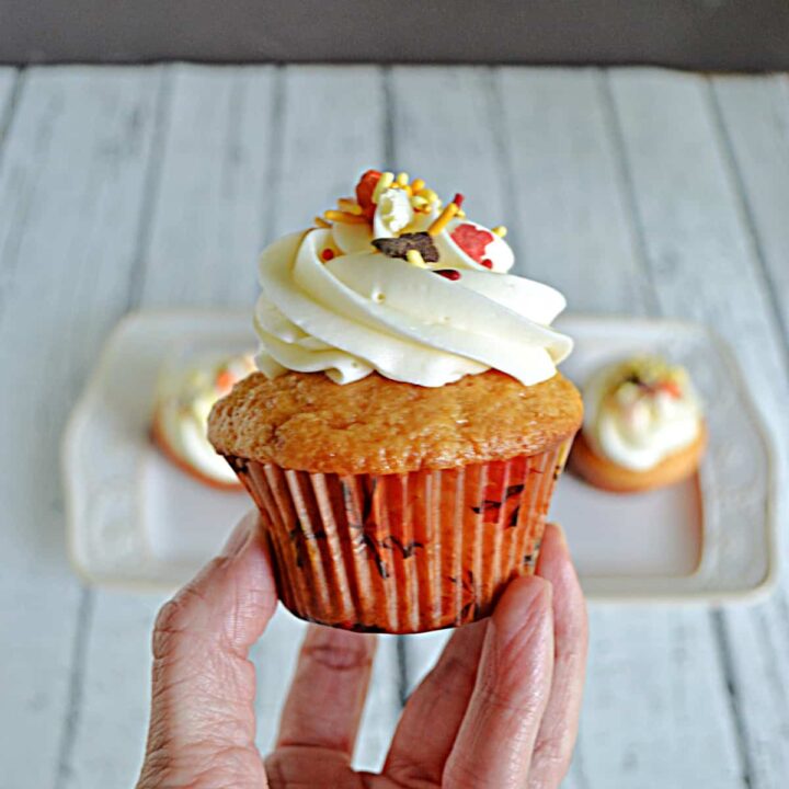 A close up of a hand holding a butterscotch cupcake with frosting and fall colored sprinkles.