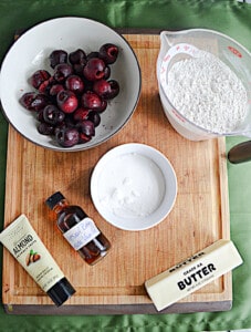 A cutting board with a bowl of cherries, a bowl of sugar, a measuring cup of flour, a stick of butter, and vanilla extract on it.