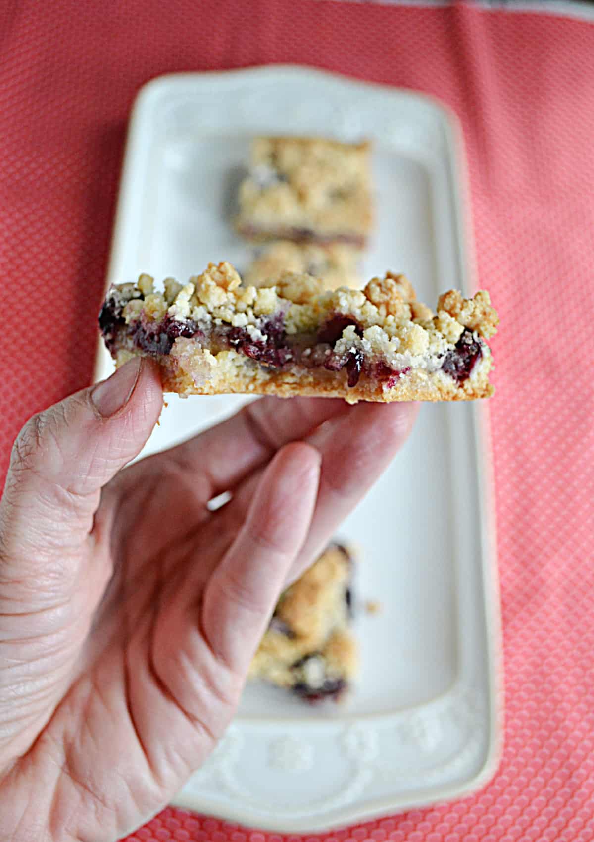 A close up of a hand holding a cherry crumble bar with a bite taken out of it.