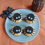 A plate of cupcakes with Oreo spiders, a toy spider on the plate, and a pumpkin behind the plate.