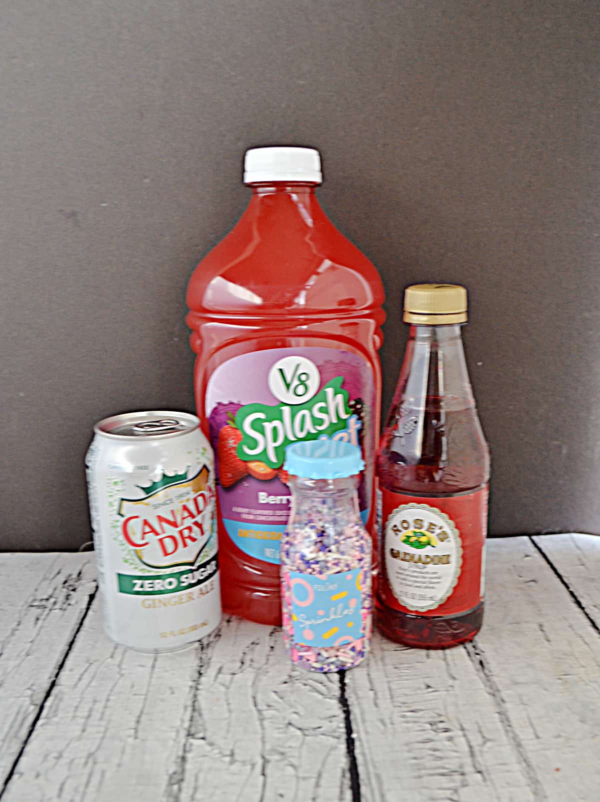 A jug of berry juice, a bottle of grenadine, a bottle of sprinkles, and a can of ginger ale.