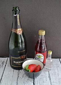 A bottle of champagne, a bowl of strawberries, a bottle of grenadine, and a bottle of vodka.