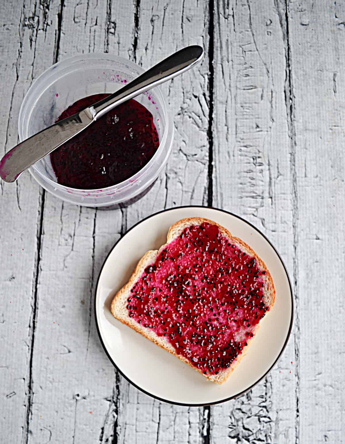 A plate with a slice of toast with jam on it and a container of jam behind it with a knife on top of it. 