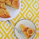 A plate with a slice of lemon cake and a fork on it with the cake behind it.