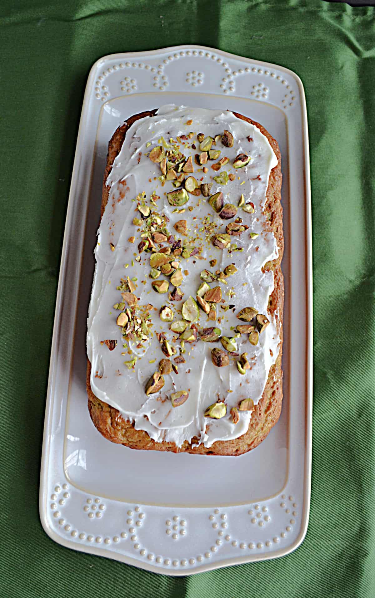 A white platter with a pistachio cake covered in a glaze and chopped pistachios on it.