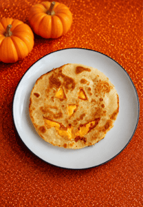 A quesadilla with a Jack-o-lantern face cut out of it on a plate with 2 pumpkins behind the plate.