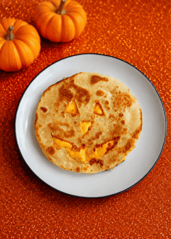A quesadilla with a Jack-o-lantern face cut out of it on a plate with 2 pumpkins behind the plate.