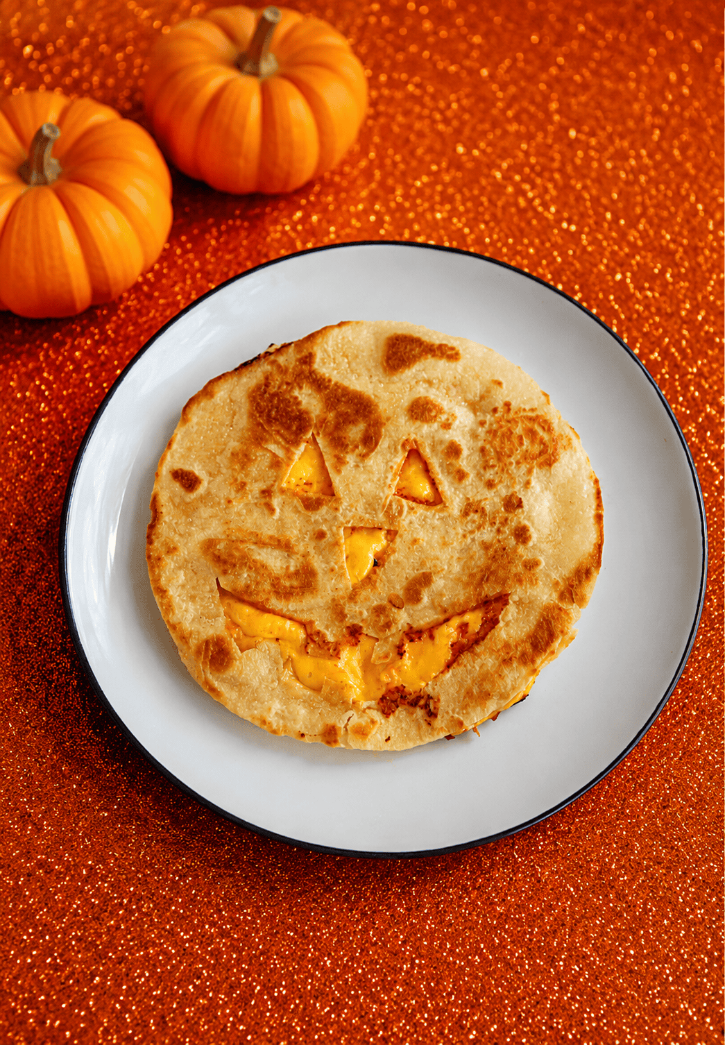 A quesadilla with a Jack-o-lantern face cut out of it on a plate with 2 pumpkins behind the plate.