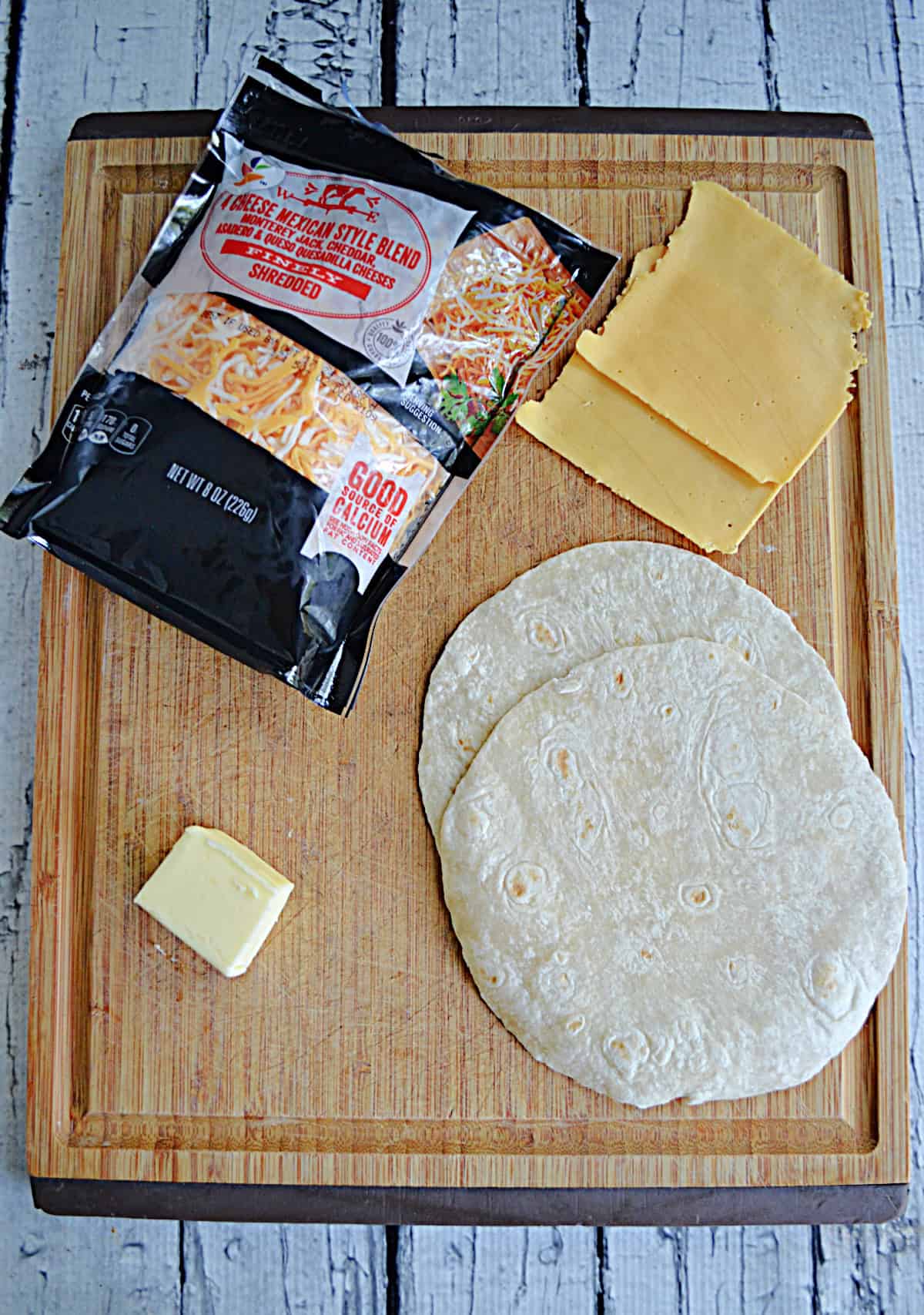 A wooden cutting board with butter, tortillas, shredded cheeses, and sandwich cheese on it.