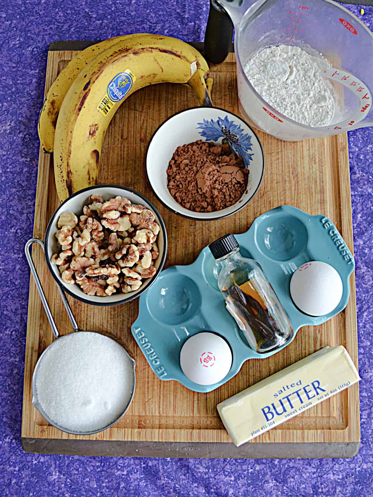 A wooden cutting board with bananas, sugar, flour, and cocoa powder.