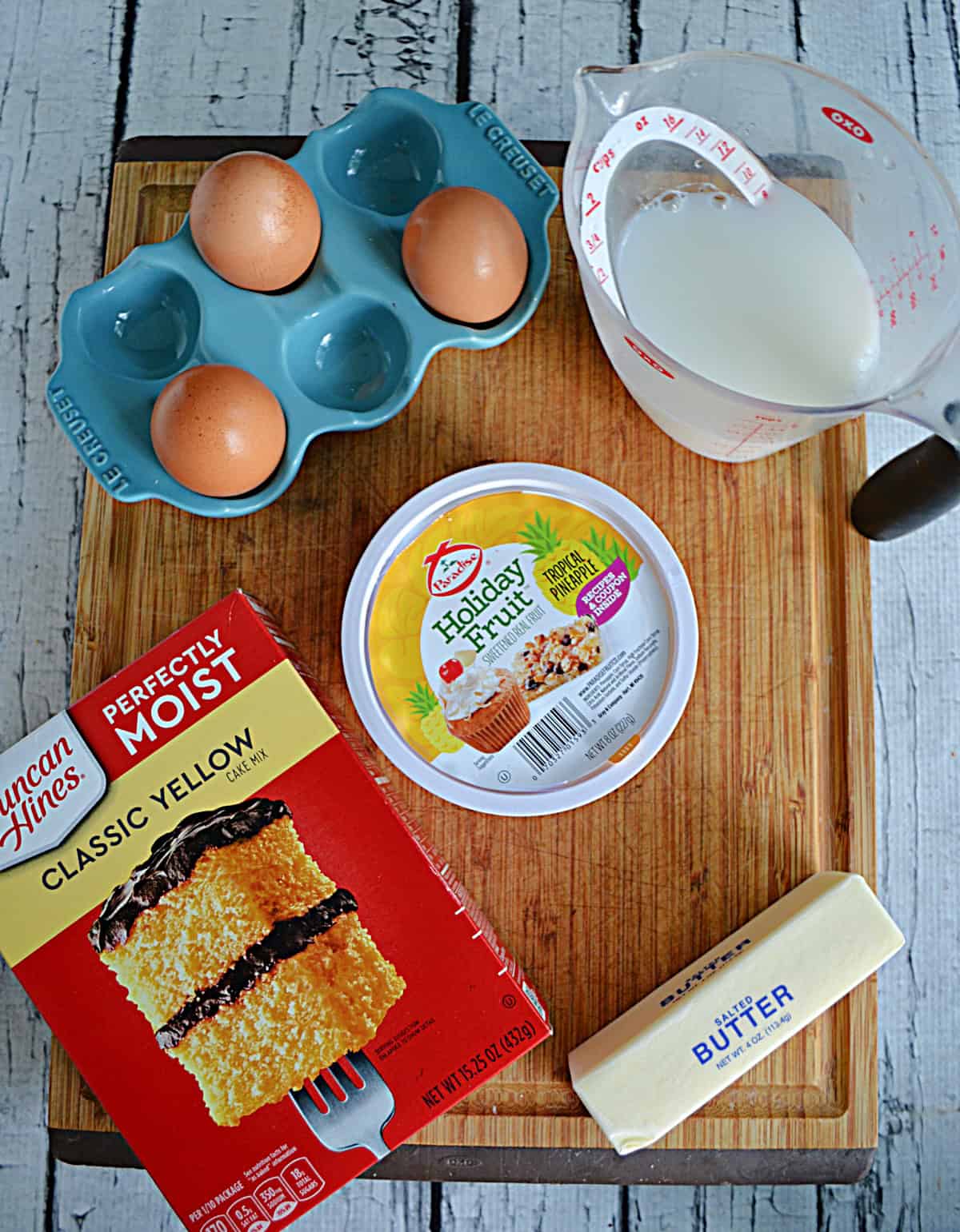 A cutting board with a box of cake mix, eggs, milk, butter, and candied pineapple on it.