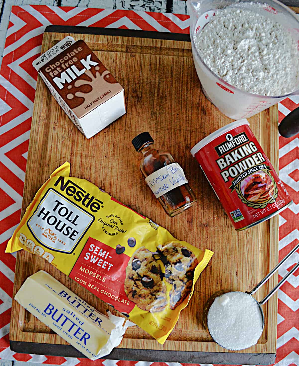 Wooden cutting board with a cup of flour, a bag of chocolate chips, vanilla, a container of chocolate milk, a stick of butter, and baking powder.