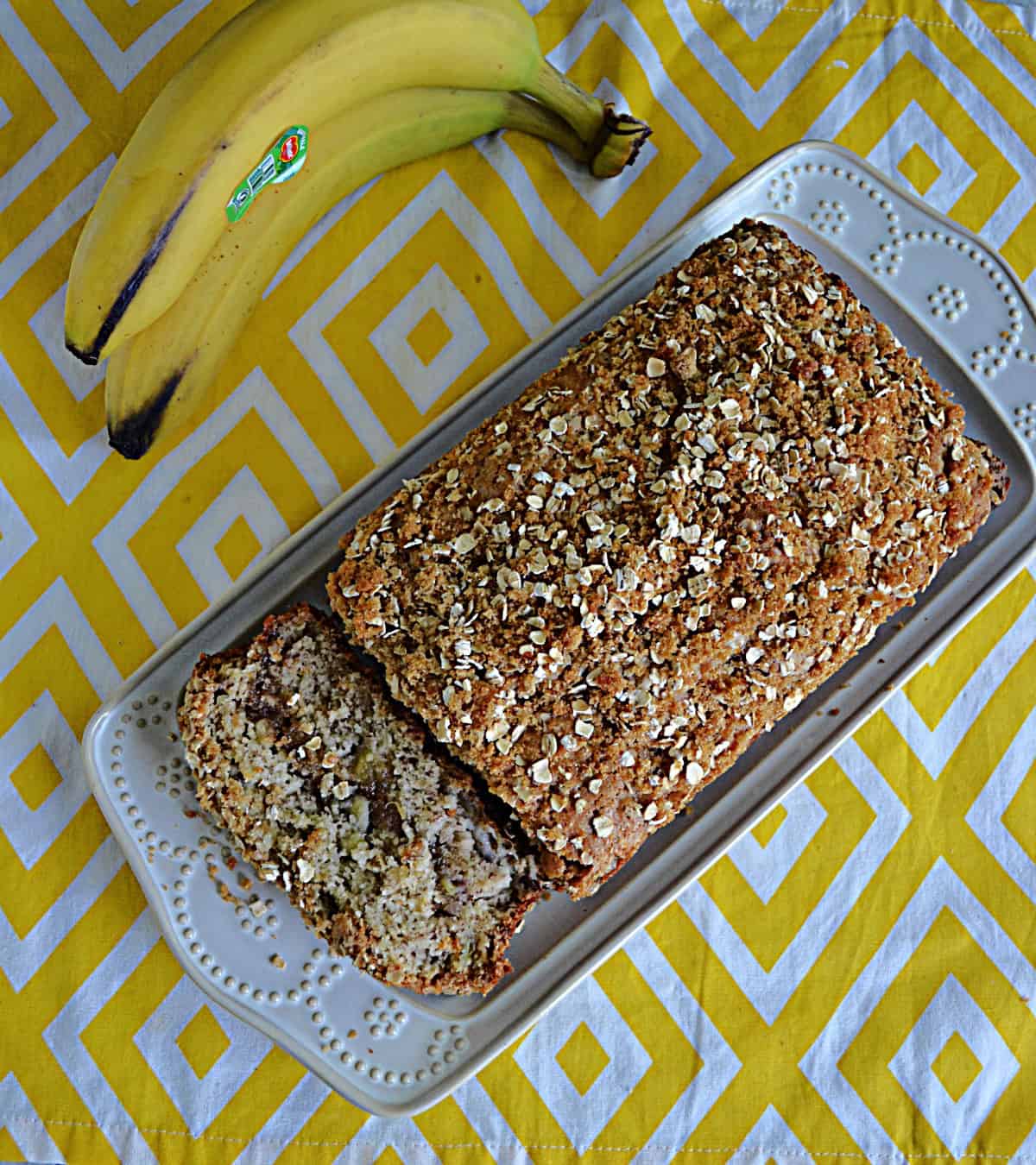 Banana Bread on a platter with a slice cut off and showing and bananas in the background.