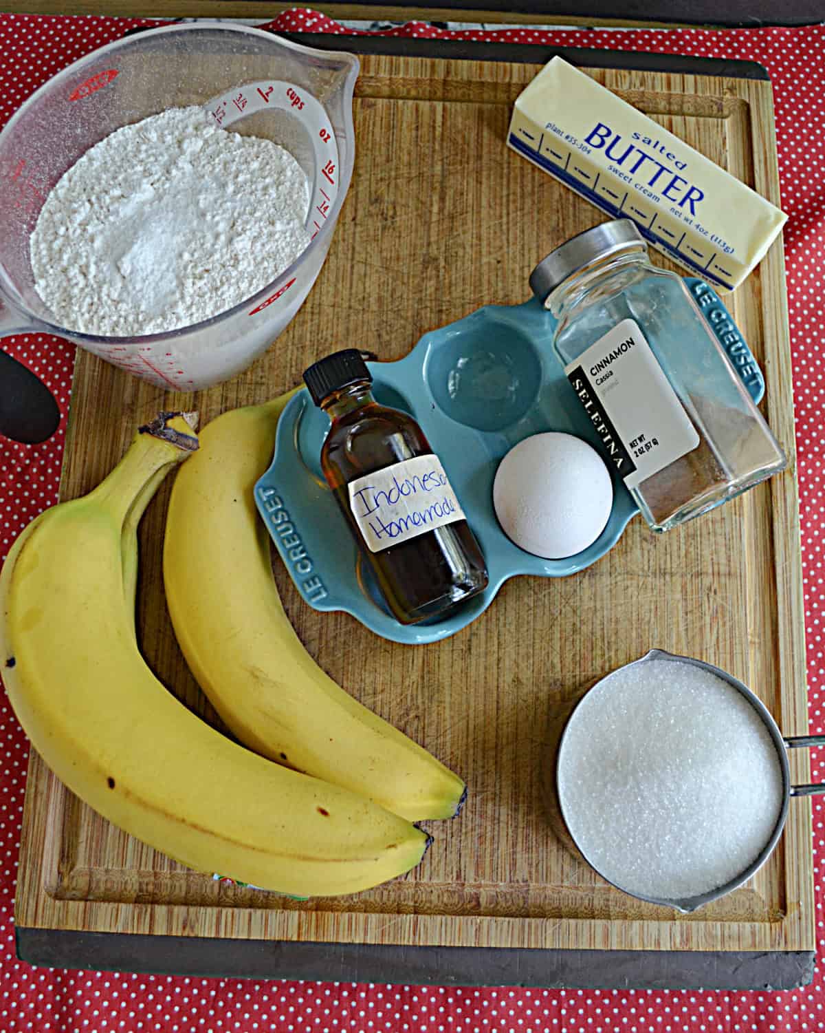 A cutting board with bananas, flour, sugar, egg, butter, vanilla, and cinnamon on it.