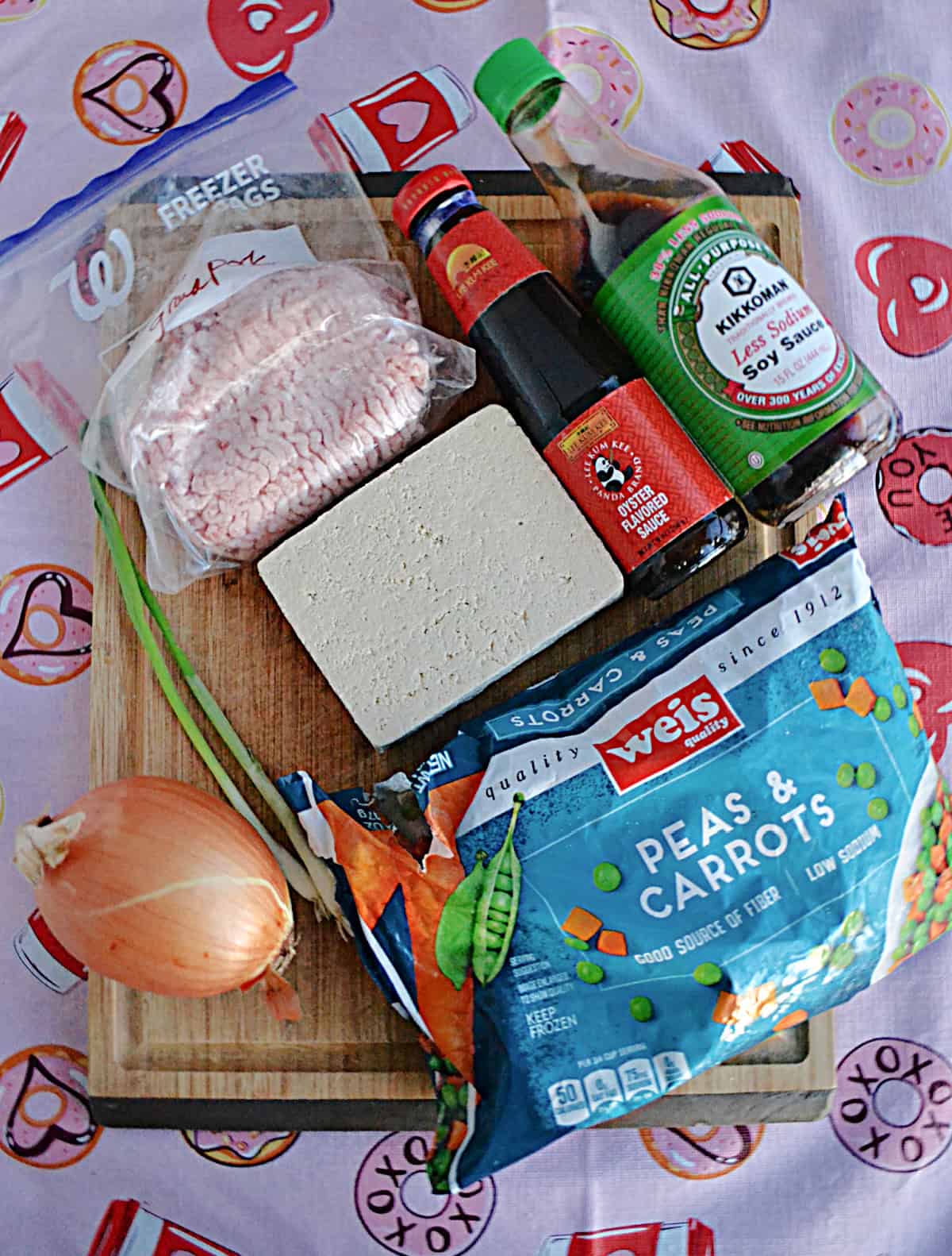 A cutting board with tofu, soy sauce, a bag of peas and carrots, an onion, and ground pork on it.
