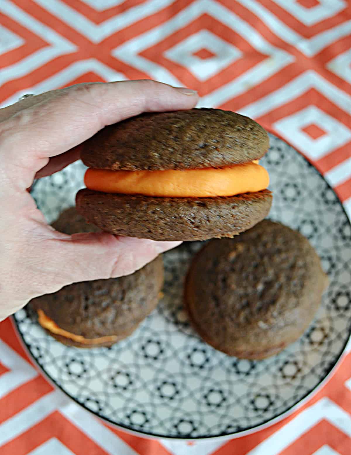 A close up of a hand holding a chocolate whoopie pie with orange filling.
