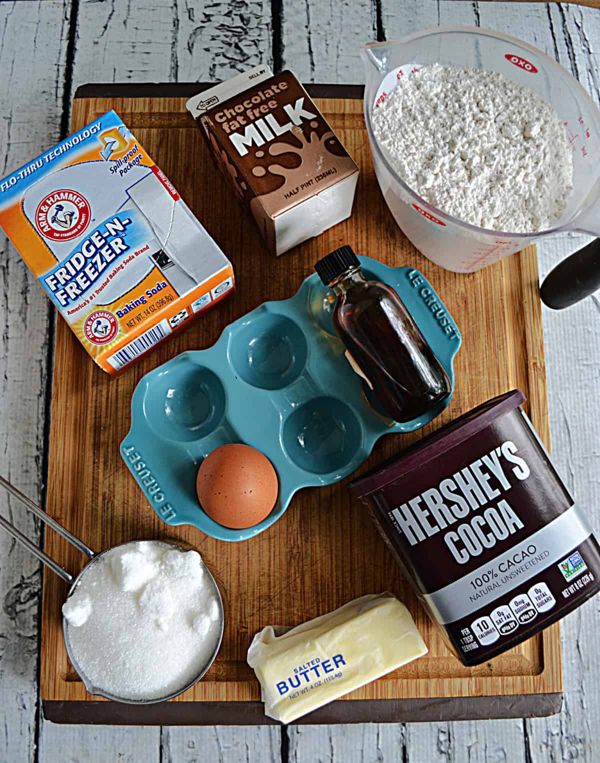 A cutting board with flour, chocolate milk, cocoa powder, sugar, egg, vanilla, and baking soda.