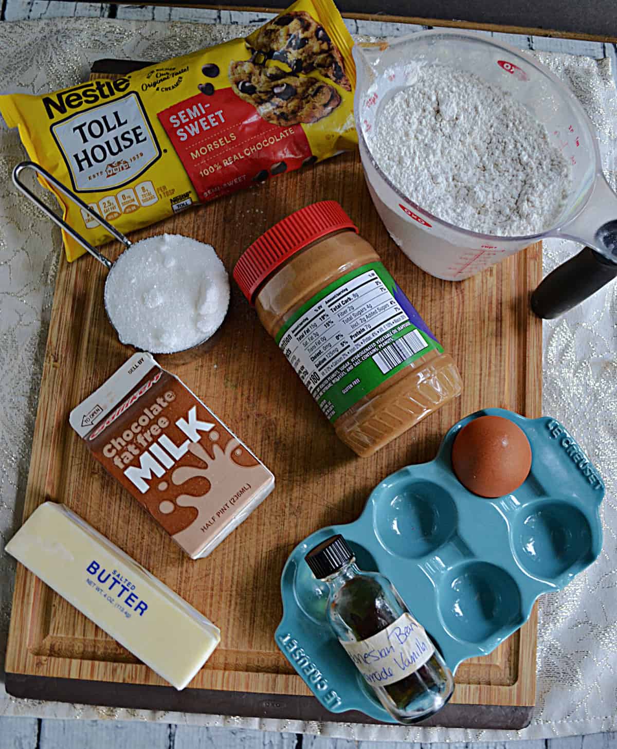 A wooden cutting board with chocolte milk, an egg, butter, a bag of chocolate chips, a cup of flour, a cup of sugar, and peanut butter.