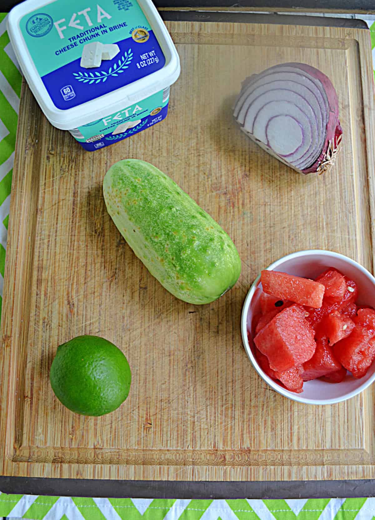 Wooden cutting board with feta cheese, cucumber, lime, onion, and watermelon on it.