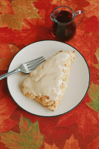 A plate with a frosted maple scone and fork with a carafe of syrup in the background.