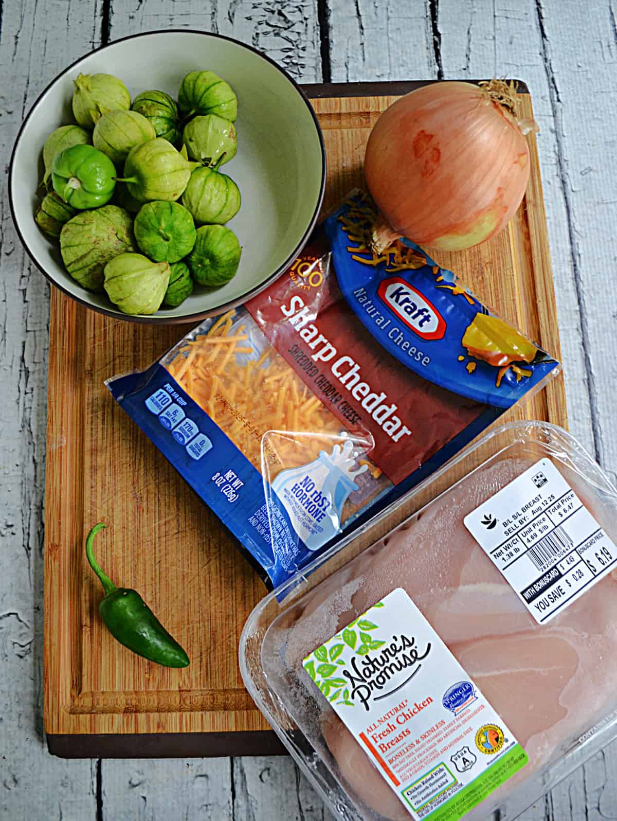 A cutting board with a bowl of tomatillos, a bag of cheese, an onion, a jalapeno, and a container or chicken on it.