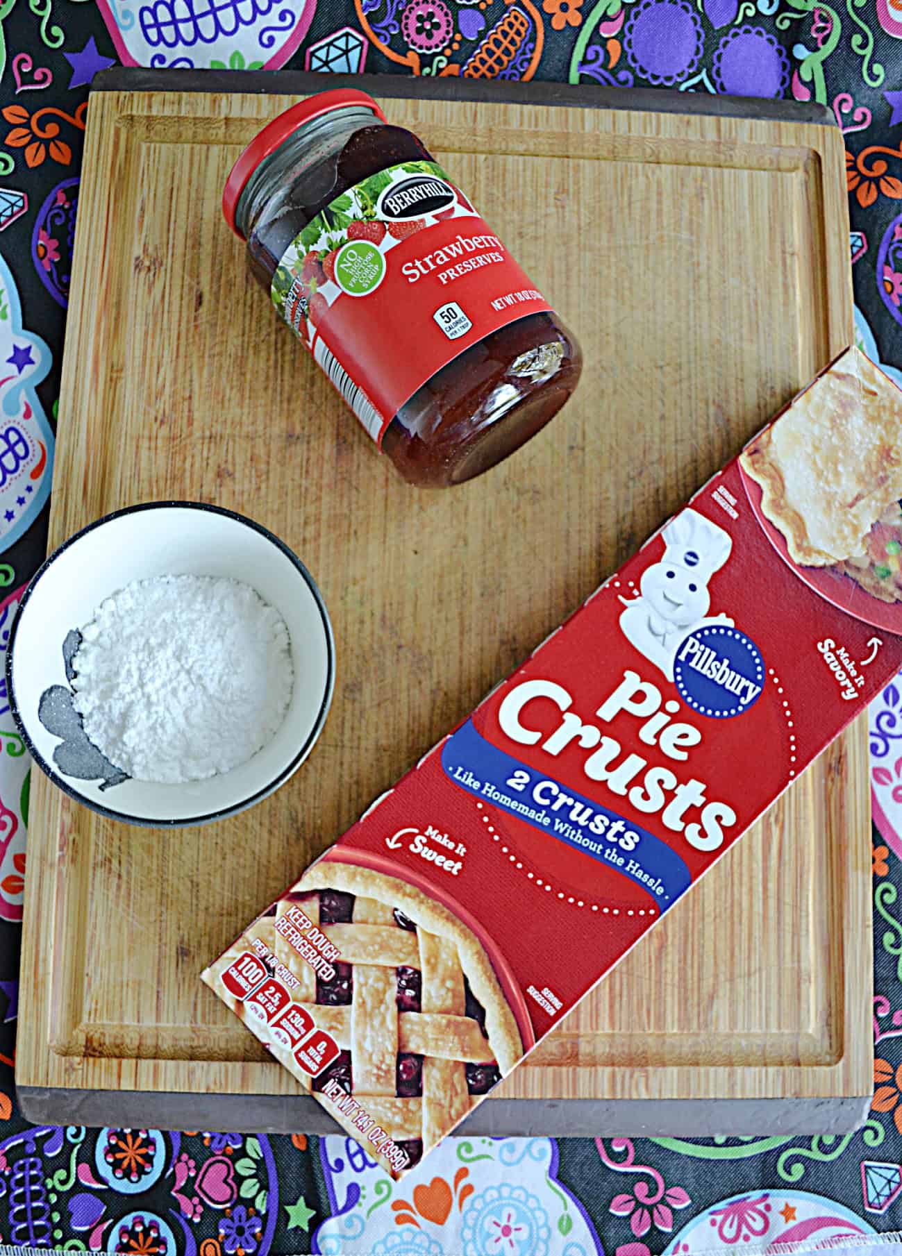 A wooden cutting board with a box of pie crusts, a bowl of powdered sugar, and a jar of strawberry jam on it.