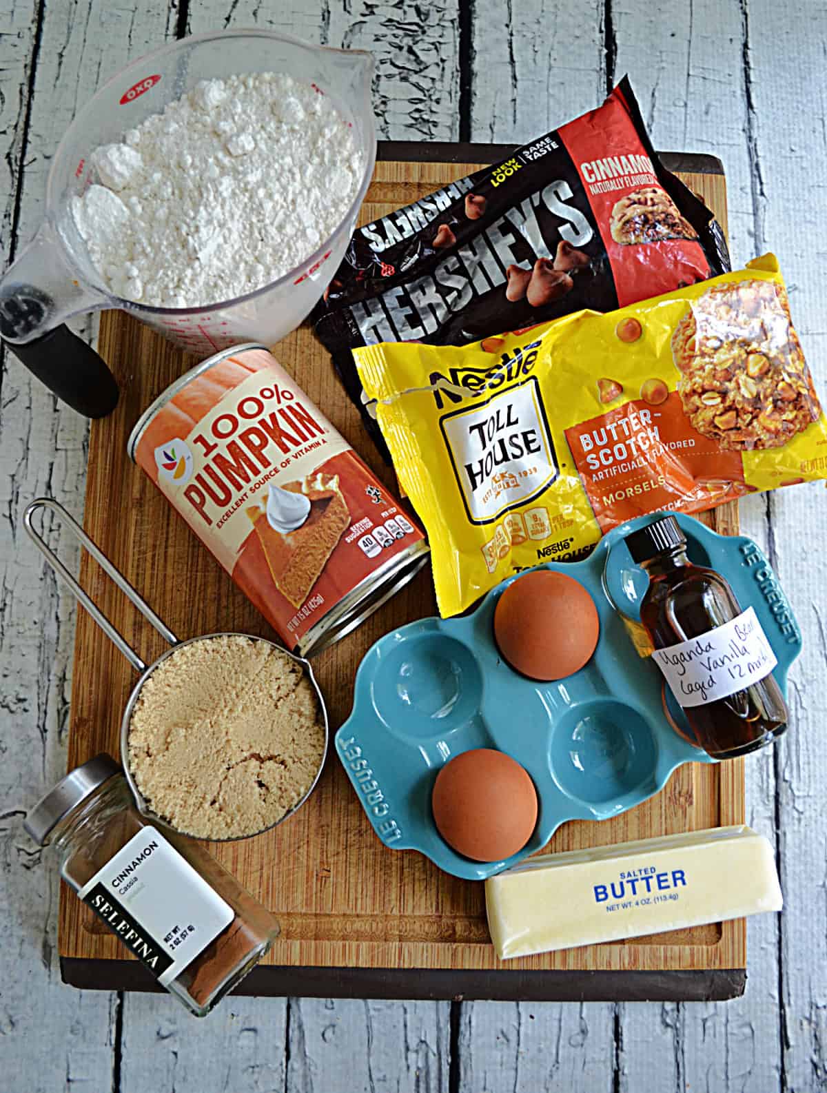 A cutting board with a cup of flour, brown sugar, eggs, butter, butterscotch chips, cinnamon chips, and pumpkin on it.