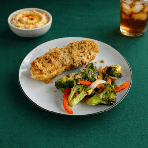A plate with a piece of chicken, broccoli. and peppers on it with a bowl of hummus and a glass of iced tea in the background.