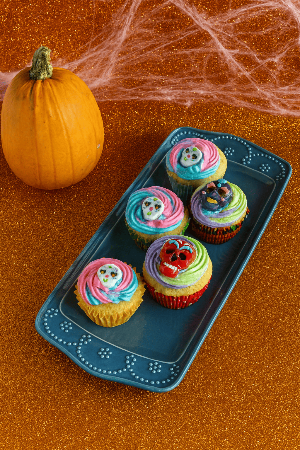 A platter of cupcakes with sugar skulls on them with spiderwebs and a pumpkin in the background.
